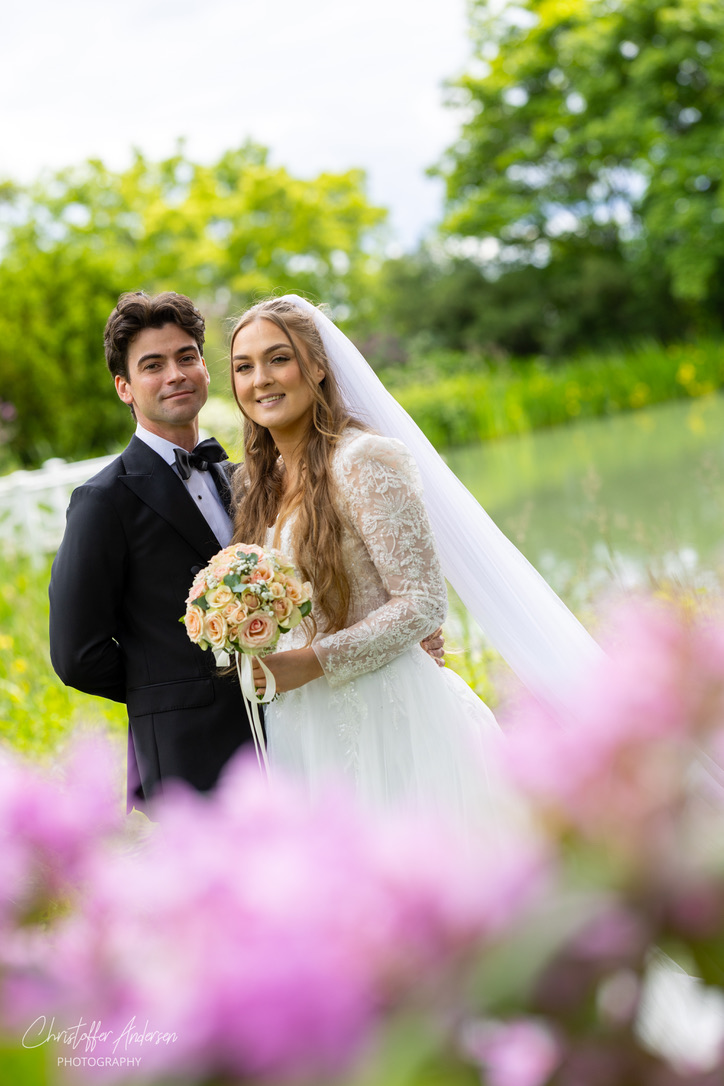Brudepar ved vann med rosa blomster i forgrunnen